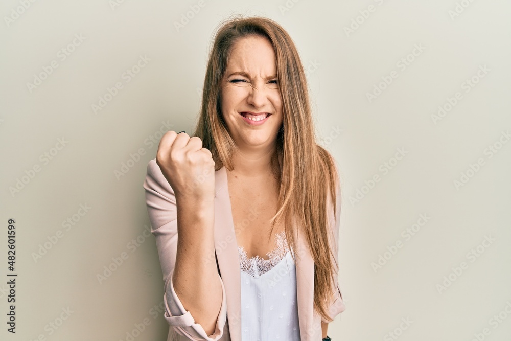 Young blonde woman wearing business jacket and glasses angry and mad raising fist frustrated and furious while shouting with anger. rage and aggressive concept.