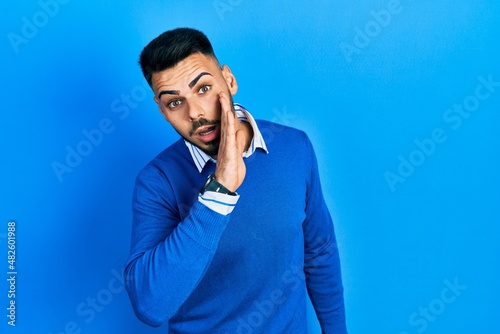 Young hispanic man with beard wearing casual blue sweater hand on mouth telling secret rumor, whispering malicious talk conversation