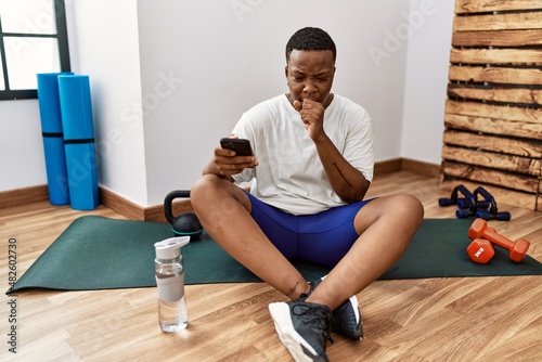 Young african man sitting on training mat at the gym using smartphone feeling unwell and coughing as symptom for cold or bronchitis. health care concept.