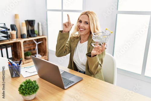 Beautiful blonde woman doing online shopping holding supermarket cart at the office surprised with an idea or question pointing finger with happy face, number one photo