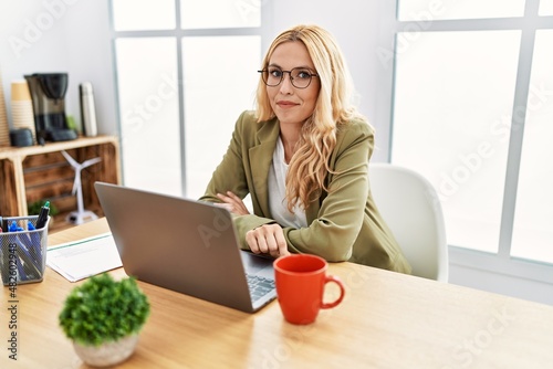 Beautiful blonde woman working at the office with laptop smiling looking to the side and staring away thinking.