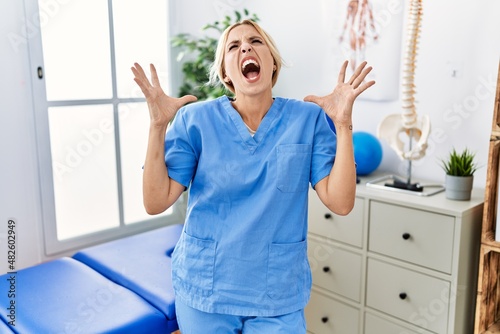 Beautiful blonde physiotherapist woman working at pain recovery clinic crazy and mad shouting and yelling with aggressive expression and arms raised. frustration concept.