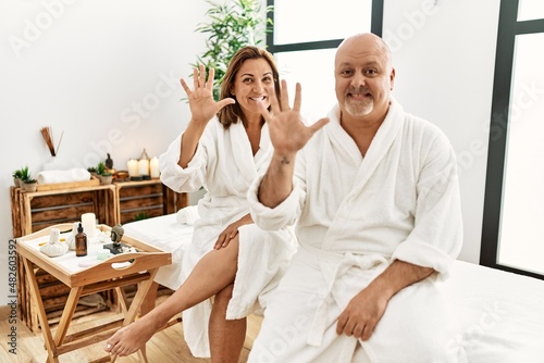 Middle age hispanic couple wearing bathrobe at wellness spa showing and pointing up with fingers number ten while smiling confident and happy.