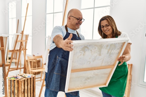 Middle age hispanic painter couple smiling happy looking canvas at art studio.