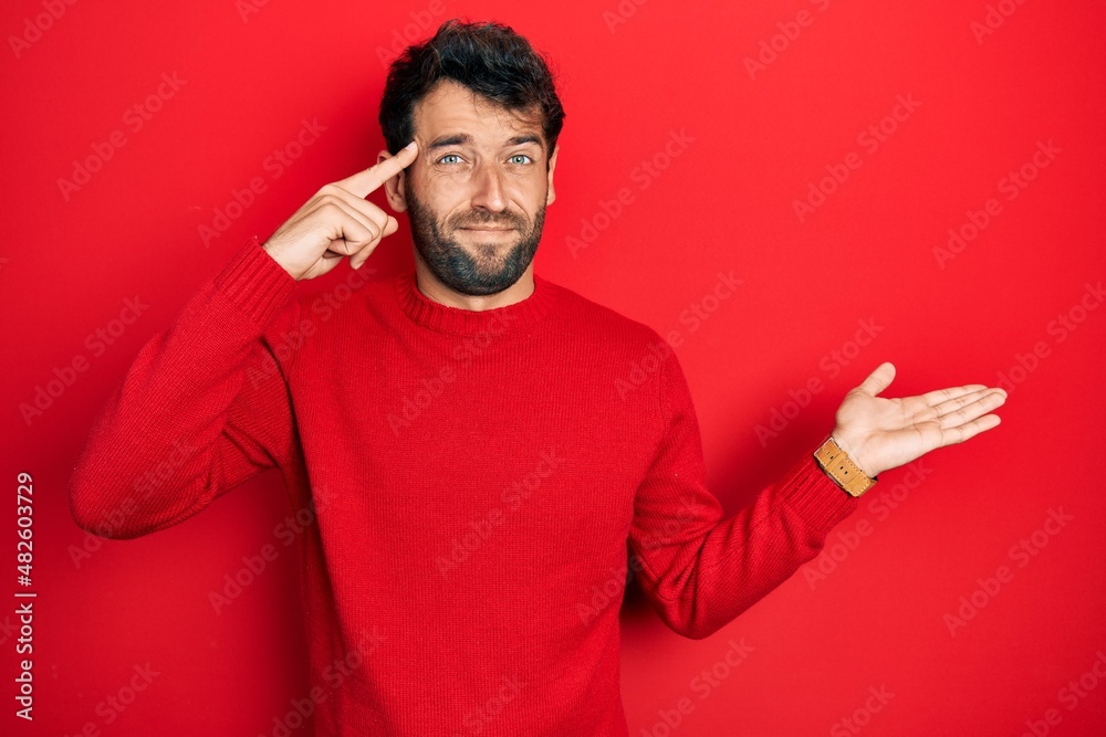 Handsome man with beard wearing casual red sweater confused and annoyed with open palm showing copy space and pointing finger to forehead. think about it. Handsome man with beard wearing casual red sweater confused and annoyed with open palm showing copy space and pointing finger to forehead. think about it.