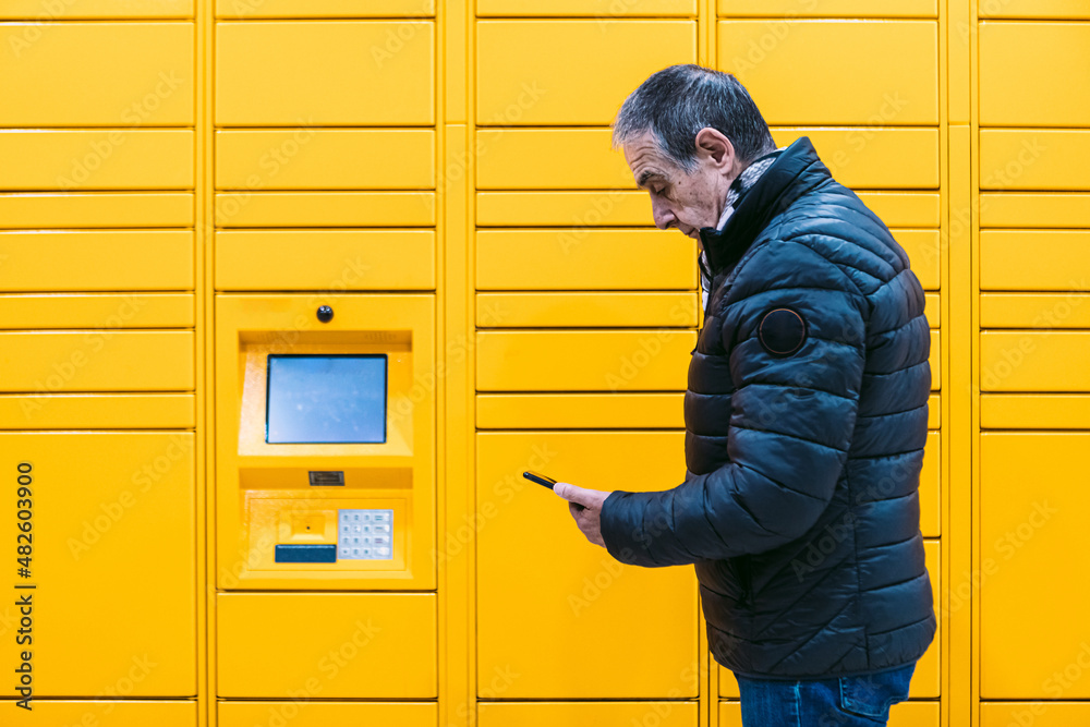 Man with gray coat looks at his cell phone to pick up a package from the yellow locker. Messaging concept, compare online, e-commerce and packages Man with gray coat looks at his cell phone to pick up a package from the yellow locker. Messaging concept, compare online, e-commerce and packages