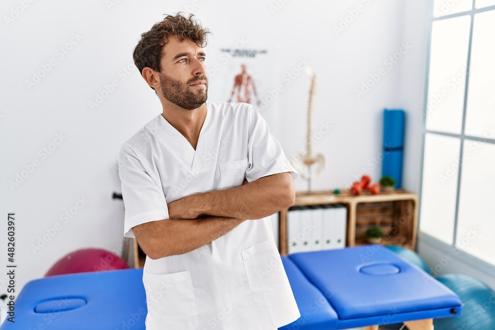 Obraz premium Young handsome physiotherapist man working at pain recovery clinic looking to the side with arms crossed convinced and confident