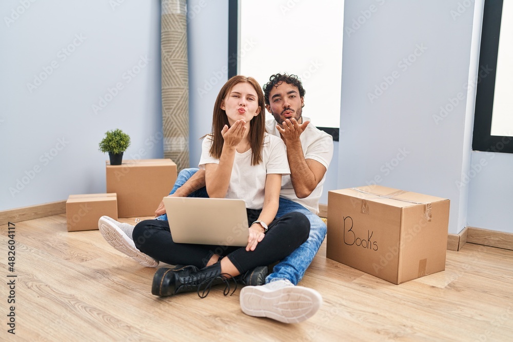 Young couple using laptop at new home looking at the camera blowing a kiss with hand on air being lovely and sexy. love expression. Young couple using laptop at new home looking at the camera blowing a kiss with hand on air being lovely and sexy. love expression.