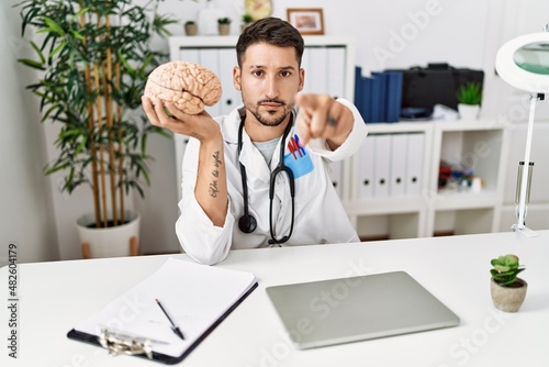 Young doctor holding brain at medical clinic pointing with finger to the camera and to you, confident gesture looking serious