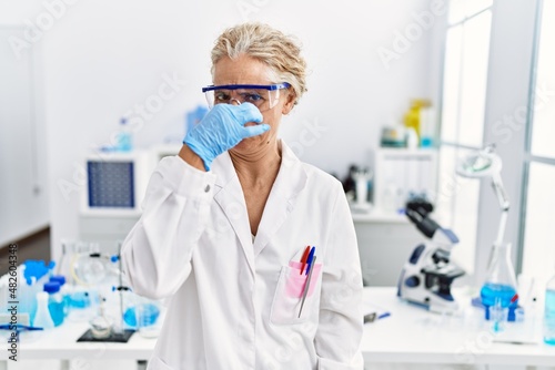 Middle age blonde woman working at scientist laboratory smelling something stinky and disgusting, intolerable smell, holding breath with fingers on nose. bad smell