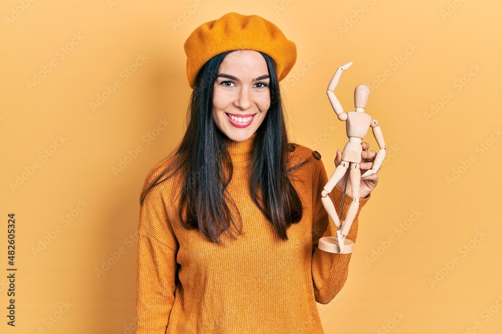Young brunette woman holding small wooden manikin wearing beret looking positive and happy standing and smiling with a confident smile showing teeth Young brunette woman holding small wooden manikin wearing beret looking positive and happy standing and smiling with a confident smile showing teeth