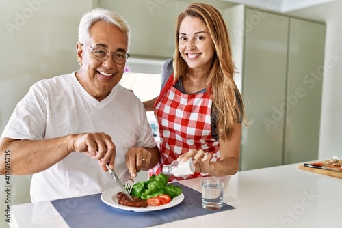 Middle age hispanic couple smiling happy eating beef with salad at the kitchen.