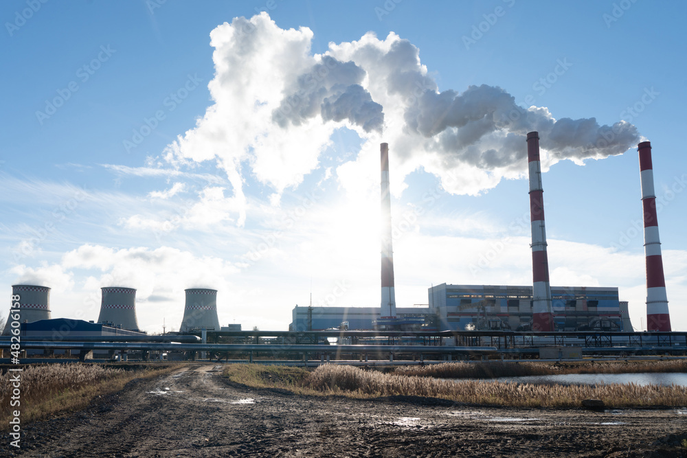 Smoking pipes of thermal power plant against blue sky. Large blue pipe releases white steam. Fuming chimneys of a factory polluting Smoking pipes of thermal power plant against blue sky. Large blue pipe releases white steam. Fuming chimneys of a factory polluting