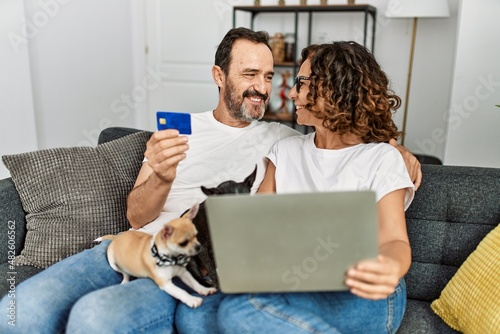 Middle age hispanic couple using laptop and credit card. Sitting on the sofa with dogs at home.