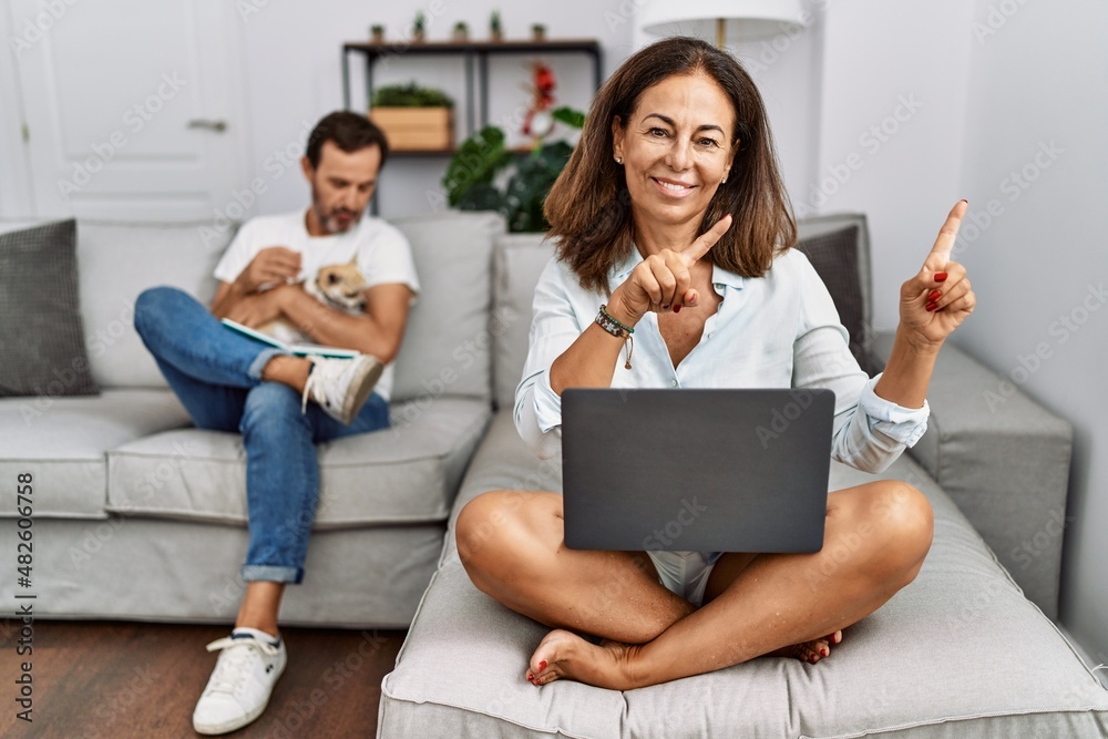 Hispanic middle age couple at home, woman using laptop smiling and looking at the camera pointing with two hands and fingers to the side. Hispanic middle age couple at home, woman using laptop smiling and looking at the camera pointing with two hands and fingers to the side.