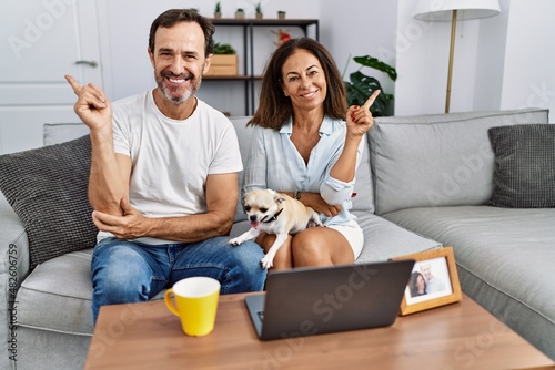 Hispanic middle age couple sitting on the sofa using computer laptop smiling happy pointing with hand and finger to the side