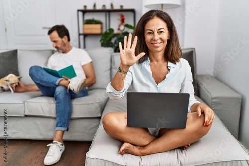 Hispanic middle age couple at home, woman using laptop showing and pointing up with fingers number five while smiling confident and happy.