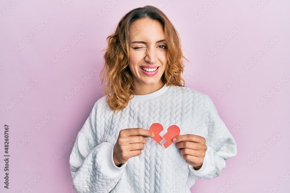 Young caucasian woman holding broken heart paper shape winking looking at the camera with sexy expression, cheerful and happy face. Young caucasian woman holding broken heart paper shape winking looking at the camera with sexy expression, cheerful and happy face.