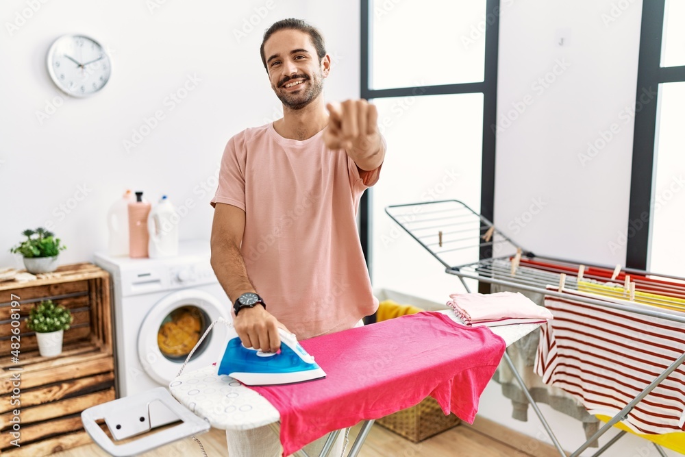 Obraz premium Young hispanic man ironing clothes at home pointing to you and the camera with fingers, smiling positive and cheerful