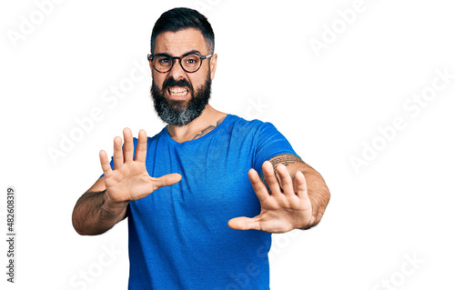 Hispanic man with beard wearing casual t shirt and glasses afraid and terrified with fear expression stop gesture with hands, shouting in shock. panic concept.