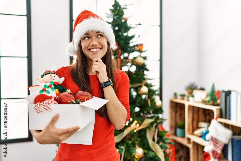 Young brunette woman standing by christmas tree holding decoration with hand on chin thinking about question, pensive expression. smiling and thoughtful face. doubt concept. Young brunette woman standing by christmas tree holding decoration with hand on chin thinking about question, pensive expression. smiling and thoughtful face. doubt concept.