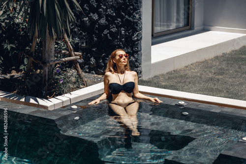 Attractive young girl wearing a beautiful black swimsuit and bright glasses is lying in a pool with clear water next to a small palm tree. Holiday concept