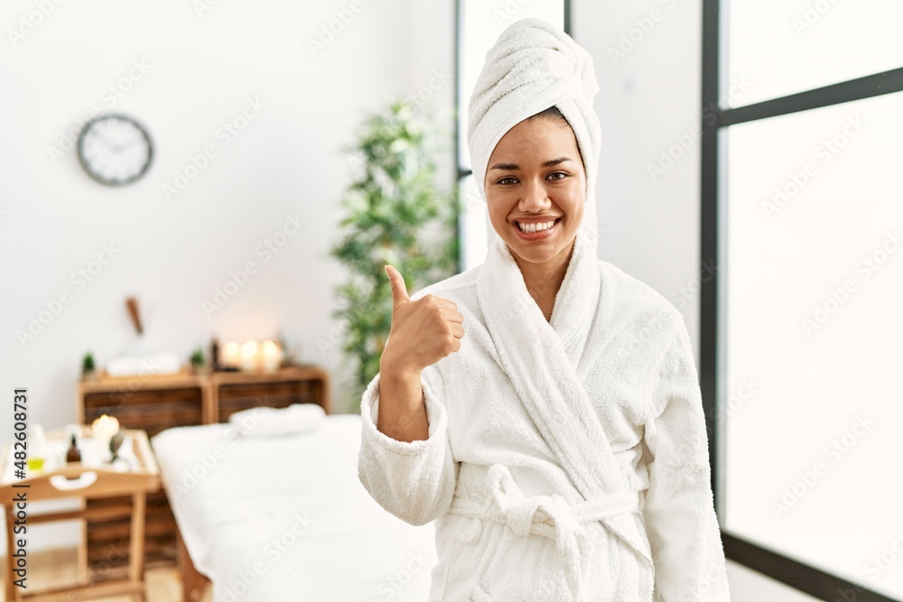 Young brunette woman wearing towel and bathrobe standing at beauty center doing happy thumbs up gesture with hand. approving expression looking at the camera showing success.
