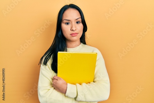 Beautiful hispanic woman with nose piercing holding book relaxed with serious expression on face. simple and natural looking at the camera.