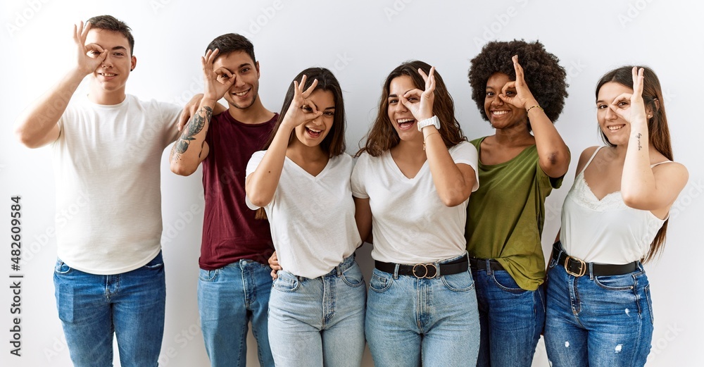 Group of young friends standing together over isolated background doing ok gesture with hand smiling, eye looking through fingers with happy face. Group of young friends standing together over isolated background doing ok gesture with hand smiling, eye looking through fingers with happy face.