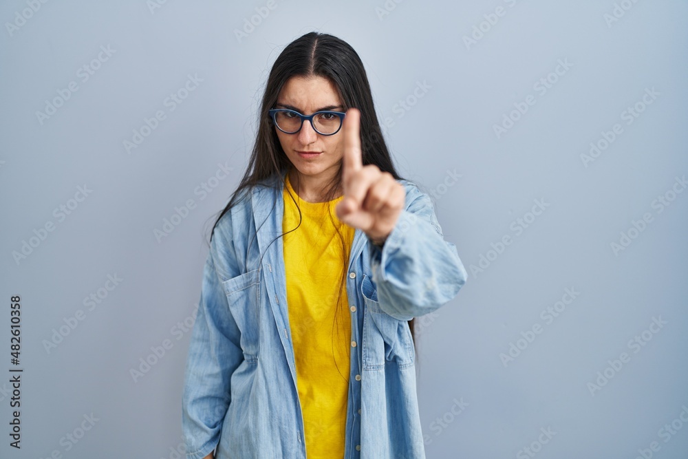 Young hispanic woman standing over blue background pointing with finger up and angry expression, showing no gesture Young hispanic woman standing over blue background pointing with finger up and angry expression, showing no gesture