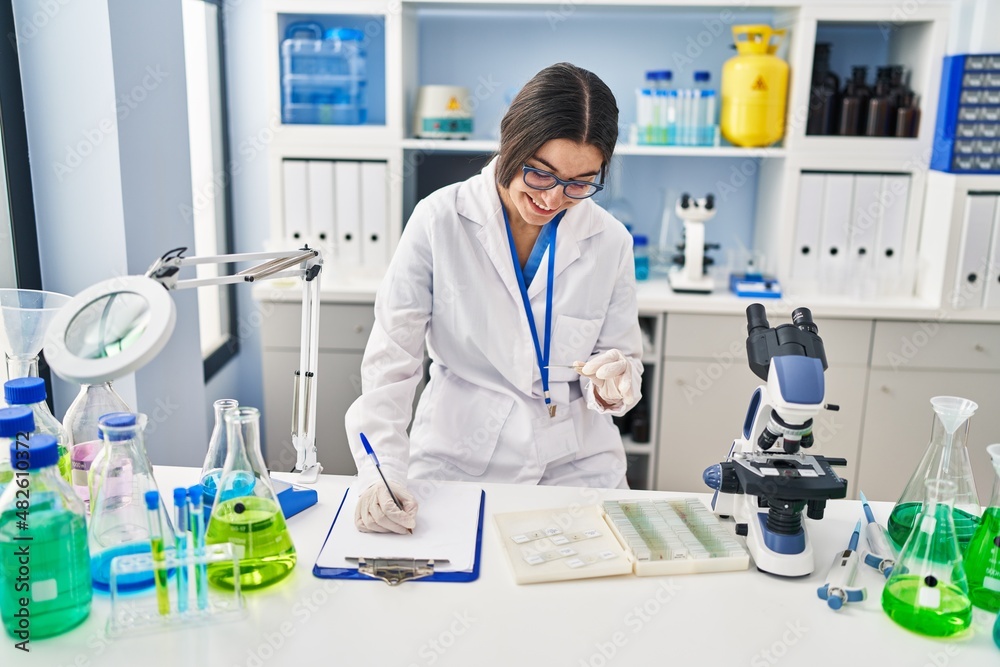Young hispanic woman wearing scientist uniform writing on clipboard holding sample at laboratory