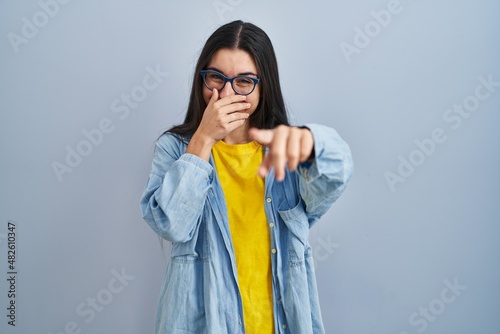 Young hispanic woman standing over blue background laughing at you, pointing finger to the camera with hand over mouth, shame expression