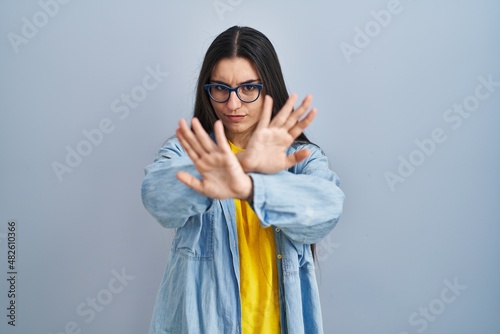 Young hispanic woman standing over blue background rejection expression crossing arms and palms doing negative sign, angry face