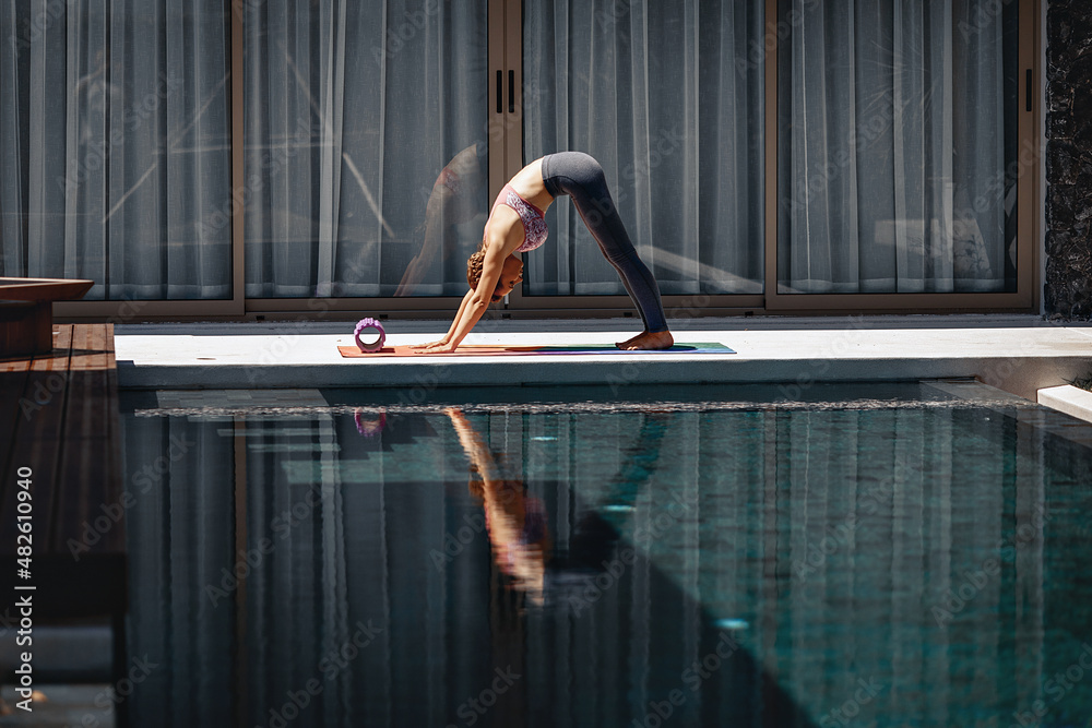Adorable skinny fit youg model doing yoga outdoors next to the pool and standing in a downward dog pose on a colorful rug next to the pool and glass doors. Health concept
