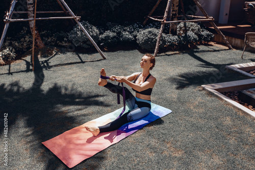 Summer outdoor activities. Young beautiful woman wearing sports tight clothes and doing yoga with yoga ribbon outdoors sitting on mat. Summer concept