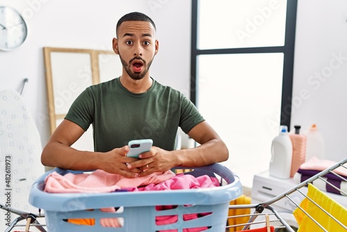 African american man doing laundry using smartphone in shock face, looking skeptical and sarcastic, surprised with open mouth
