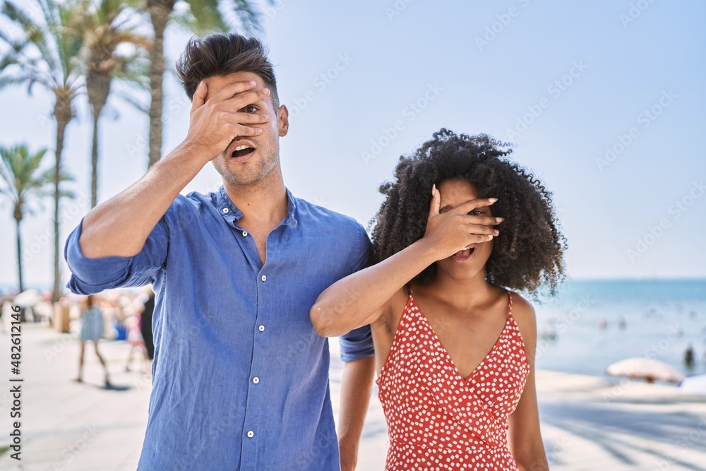 Young interracial couple outdoors on a sunny day peeking in shock covering face and eyes with hand, looking through fingers with embarrassed expression. Young interracial couple outdoors on a sunny day peeking in shock covering face and eyes with hand, looking through fingers with embarrassed expression.