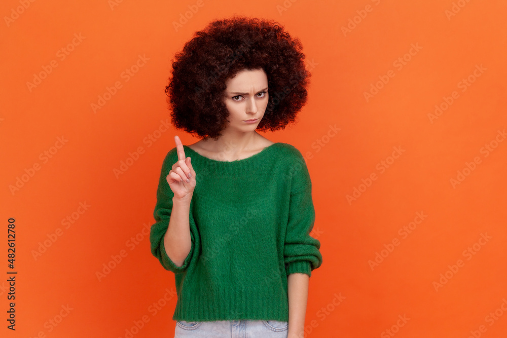 Portrait of serious woman with Afro hairstyle wearing green casual style sweater looking at camera, showing warning attention sign. Indoor studio shot isolated on orange background. - obrazy, fototapety, plakaty 