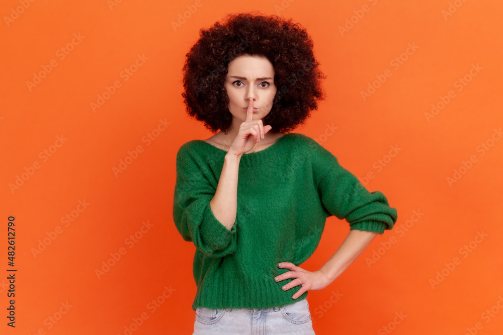 Portrait of serious woman with Afro hairstyle wearing green casual style sweater standing with finger near lips, showing silent gesture. Indoor studio shot isolated on orange background. Portrait of serious woman with Afro hairstyle wearing green casual style sweater standing with finger near lips, showing silent gesture. Indoor studio shot isolated on orange background.