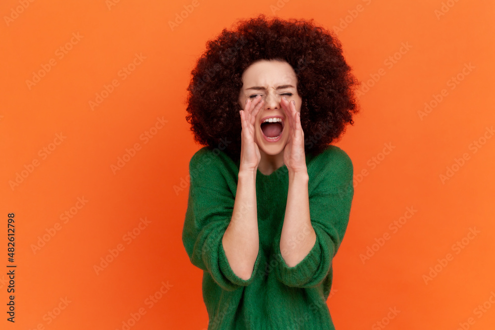 Portrait of angry yelling woman with Afro hairstyle wearing green casual style sweater keeping hands near mouth, screaming loudly. Indoor studio shot isolated on orange background. Portrait of angry yelling woman with Afro hairstyle wearing green casual style sweater keeping hands near mouth, screaming loudly. Indoor studio shot isolated on orange background.