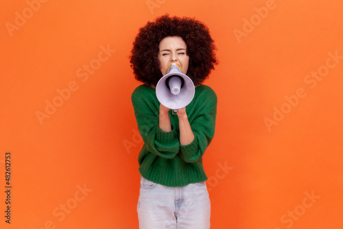 Attractive woman with Afro hairstyle wearing green casual style sweater screaming loud using megaphone, making announcement. presentation. Indoor studio shot isolated on orange background. photo