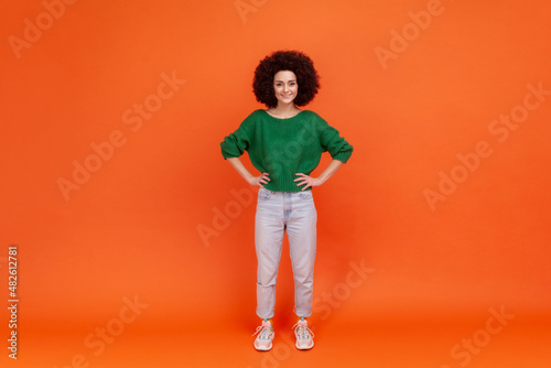 Full length portrait of positive woman with Afro hairstyle wearing green casual style sweater standing with hands on hips and toothy smile. Indoor studio shot isolated on orange background. © khosrork