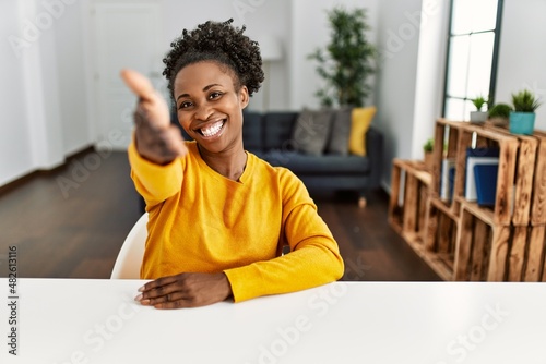 Young african american woman wearing casual clothes sitting on the table at home smiling friendly offering handshake as greeting and welcoming. successful business.