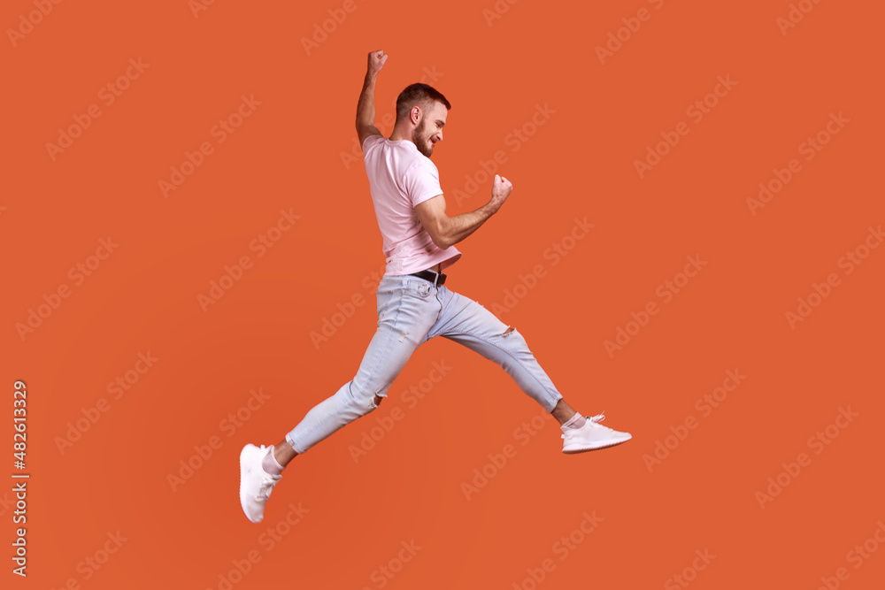 Side view portrait of happy bearded man jumping in air showing yes i did it gesture, copy space for ad, wearing pink T-shirt and jeans. Indoor studio shot isolated on orange background. Side view portrait of happy bearded man jumping in air showing yes i did it gesture, copy space for ad, wearing pink T-shirt and jeans. Indoor studio shot isolated on orange background.