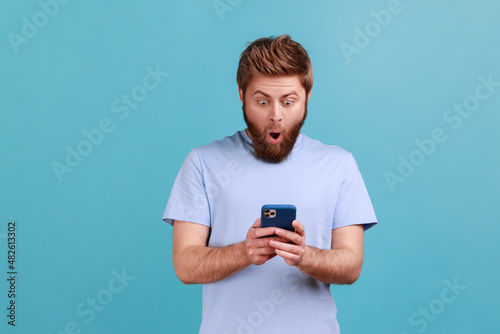 Portrait of astonished surprised young adult bearded man standing, using smartphone and watching video with amazed face and open mouth. Indoor studio shot isolated on blue background. © khosrork