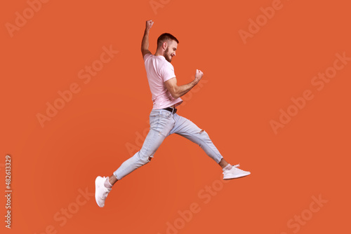 Side view portrait of happy bearded man jumping in air showing yes i did it gesture, copy space for ad, wearing pink T-shirt and jeans. Indoor studio shot isolated on orange background.