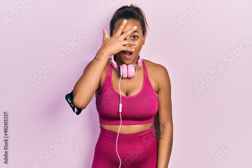 Young african american girl wearing gym clothes and using headphones peeking in shock covering face and eyes with hand, looking through fingers with embarrassed expression.