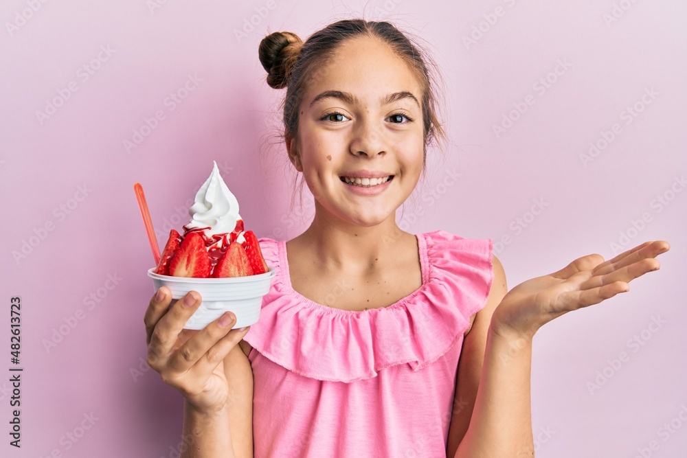 Beautiful brunette little girl eating strawberry ice cream celebrating achievement with happy smile and winner expression with raised hand