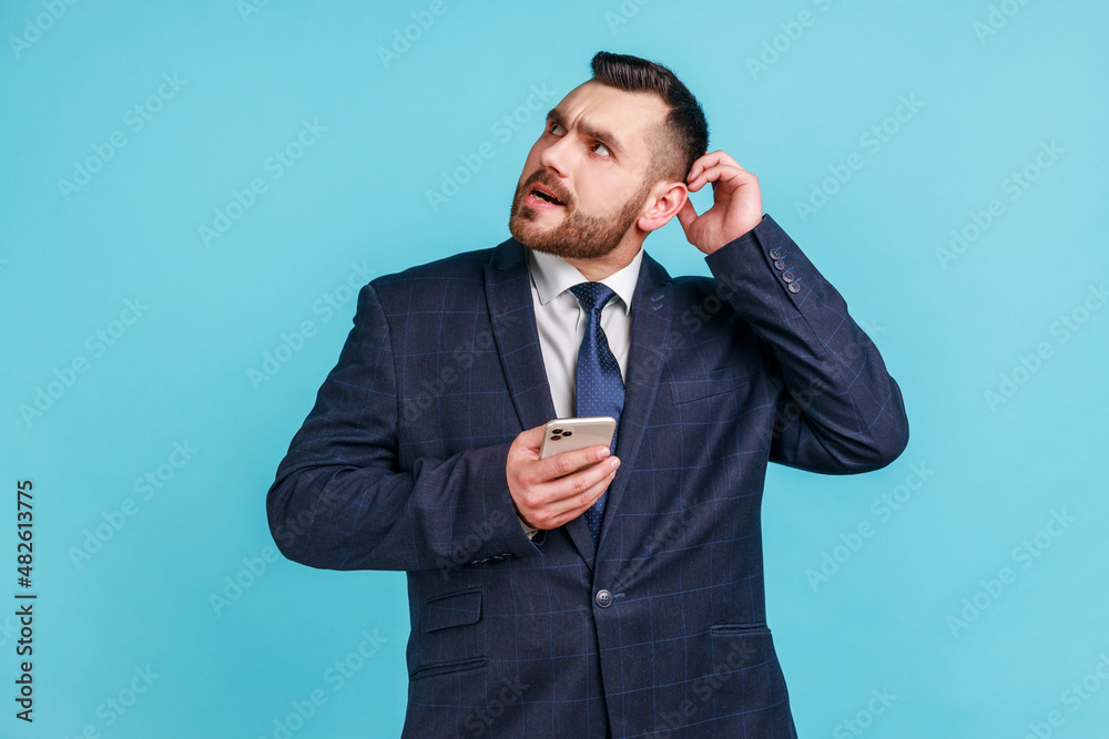 Businessman wearing official style suit scratching head holding smartphone in hand, contemplating about software updating, choosing suitable tariffs. Indoor studio shot isolated on blue background. Businessman wearing official style suit scratching head holding smartphone in hand, contemplating about software updating, choosing suitable tariffs. Indoor studio shot isolated on blue background.