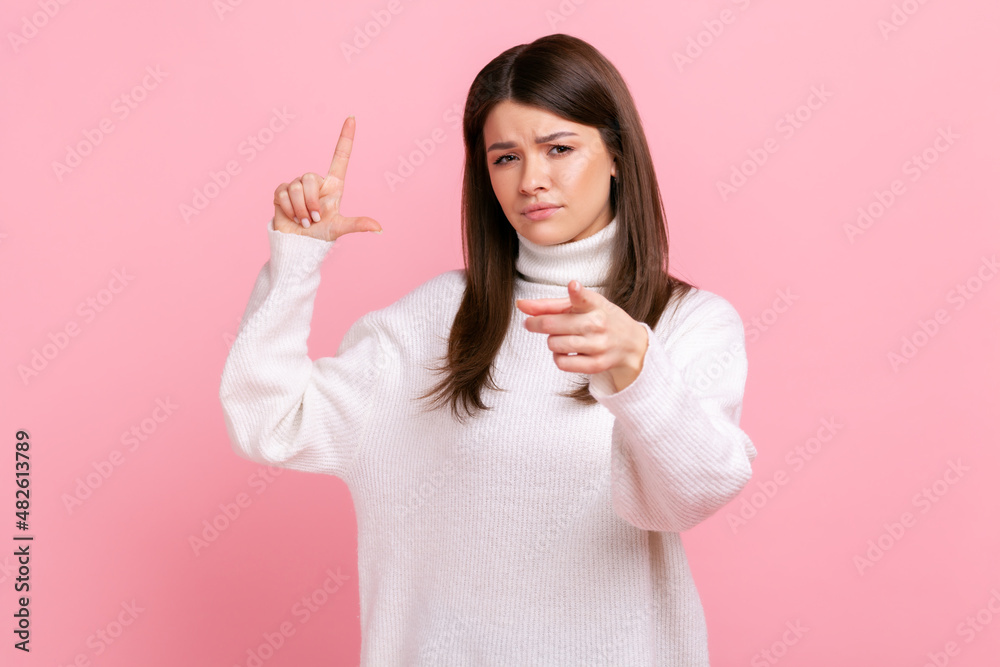 Woman expressing disrespect, showing L finger sign to camera, loser gesture, accusing for failure, wearing white casual style sweater. Indoor studio shot isolated on pink background. Woman expressing disrespect, showing L finger sign to camera, loser gesture, accusing for failure, wearing white casual style sweater. Indoor studio shot isolated on pink background.